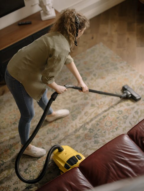 Close-up of a person holding a yellow commercial carpet cleaning machine with a black nozzle attachment, positioned on a patterned carpet in a room with soft lighting. The machine's surface is clean a