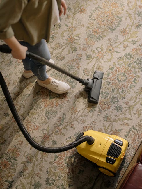 A person is vacuuming a traditional patterned area rug with a yellow and black vacuum cleaner in a well-lit room, possibly a living room or hallway. The individual's legs and feet are visible, wearing